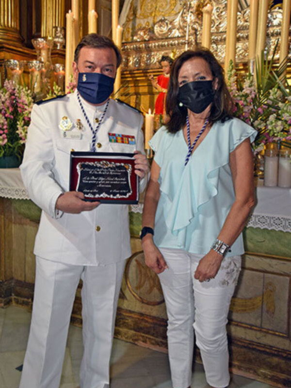 Un hombre en uniforme y una mujer en blanco posan con un certificado en un ambiente festivo y decorado con velas y flores.