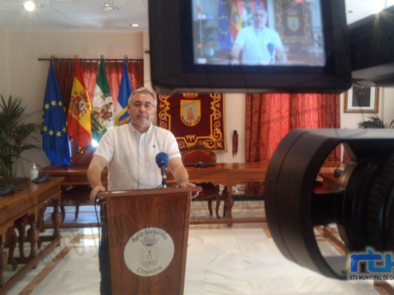 Un hombre habla desde un podio en un salón oficial con banderas y un escudo, mientras se graba con una cámara de televisión.