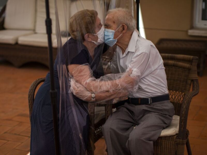 Dos ancianos se abrazan en una sala con muebles de madera y un suelo de baldosas marrones, separados por una pantalla transparente para mantener la distancia social.