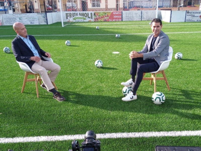 Dos hombres sentados en sillas blancas en un campo de fútbol, rodeados de balones de fútbol. El campo está rodeado por anuncios y una cámara de televisión está enfocada en ellos.