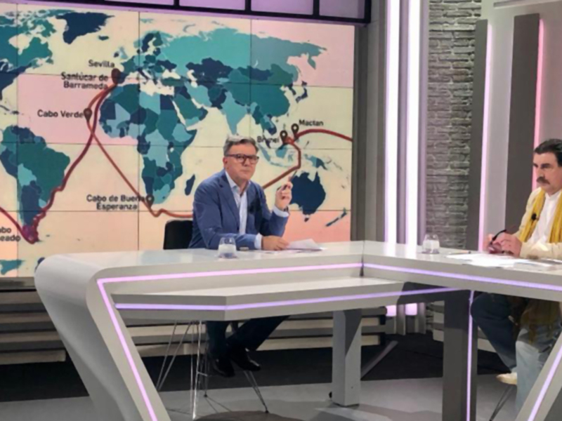 Dos personas sentadas en un estudio de televisión, uno con una camisa azul y el otro con una chaqueta beige y una bufanda amarilla, frente a un mapa del mundo que muestra varias ubicaciones geográficas.
