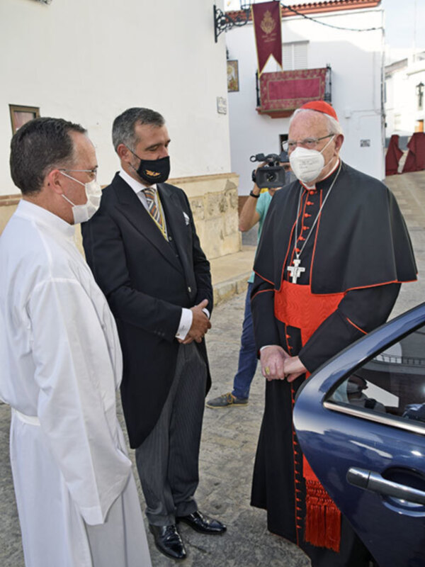 Un cardenal y otros dos hombres en trajes discuten cerca de un coche en una calle con edificios blancos.