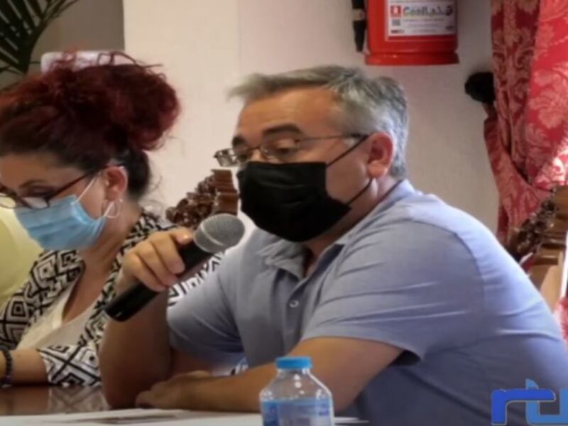 Persona hablando en un evento con microfono y máscara facial, en un ambiente formal con otro participante.
