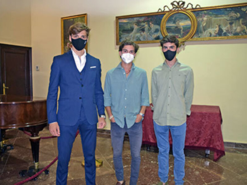 Tres personas con máscaras faciales posan juntas en un salón con pinturas antiguas y un piano de cola.