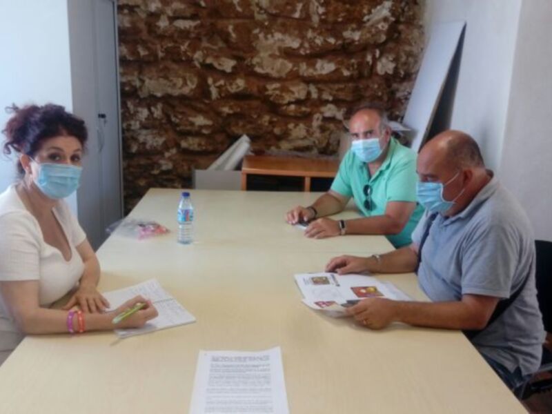 Tres personas discuten en una mesa con documentos y mascarillas, probablemente en un contexto de trabajo o reunión.
