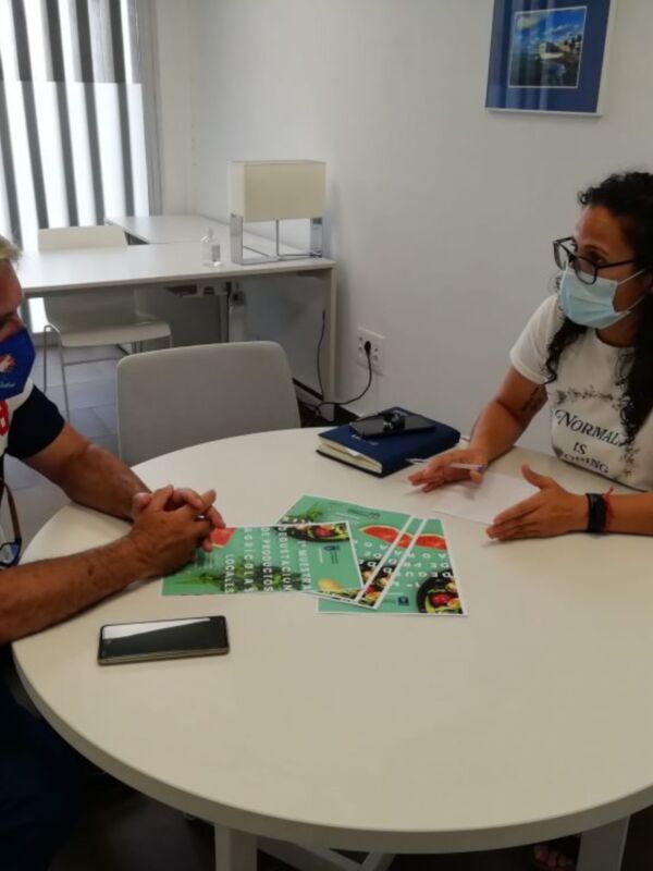Dos personas sentadas en una mesa, una con una camiseta blanca y la otra con una camiseta azul, ambos con mascarillas, sosteniendo un folleto con información sobre la salud y la prevención del coronavirus.