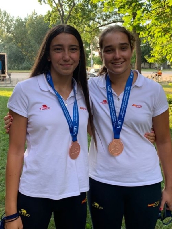Dos atletas posan orgullosas con sus medallas de bronce en un entorno verde y soleado. Ambas llevan camisetas blancas con logotipos de equipos deportivos y medallas azules con insignias de competiciones.