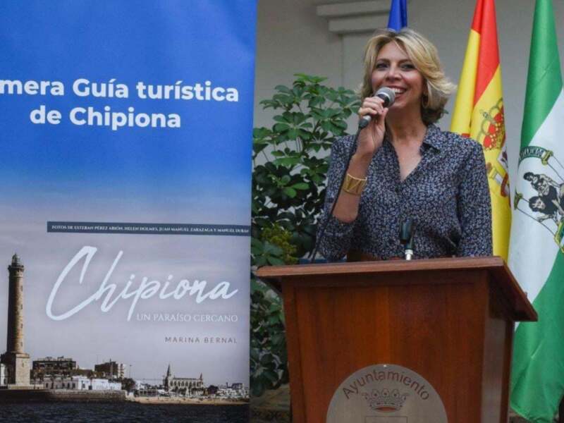 Presentación de la Primera Guía Turística de Chipiona en un evento oficial con banderas y un lighthouse en el fondo.