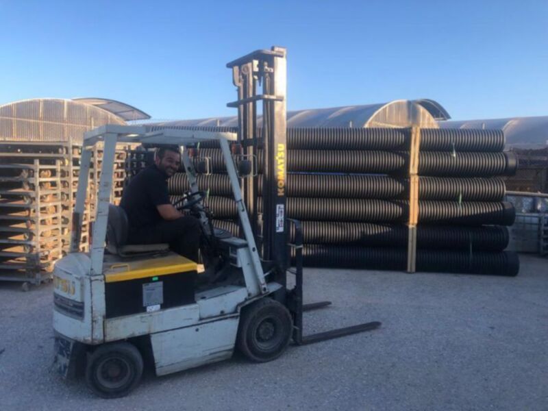 Un trabajador maneja un carretilla elevadora en un almacén, con tubos de plástico en el fondo. La imagen muestra un entorno industrial con estructuras metálicas y un cielo azul claro.