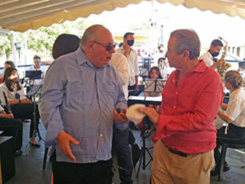 Dos hombres conversan en un evento al aire libre bajo una carpa, con otros asistentes y stands en el fondo.