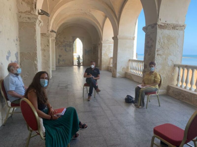 Tres personas sentadas en una terraza con arcos, con máscaras faciales, posiblemente en una reunión o conferencia.