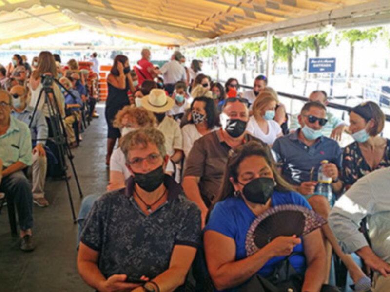 Gente reunida en un evento al aire libre, algunos con máscaras faciales, bajo una estructura de sombra.
