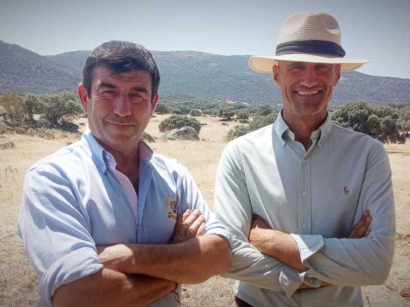 Dos hombres sonrientes con el paisaje de montañas y pasto seco en el fondo.