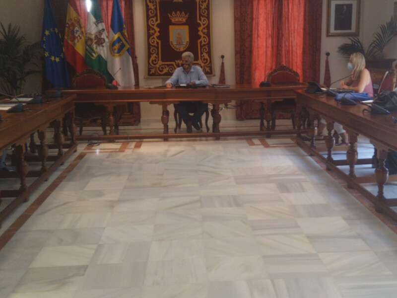 Sala de reuniones con personas sentadas en mesas, banderas y un escudo en el fondo.
