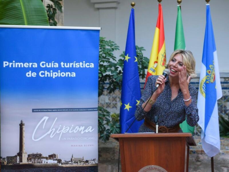 Primera Guía turística de Chipiona, con una presentación en el escenario frente a banderas europeas y un cartel con la imagen de la ciudad.