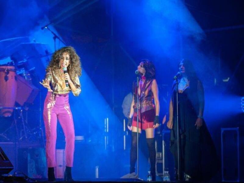 Una cantante en un concierto, con dos músicos detrás, iluminada por luces azules y con un escenario de fondo.