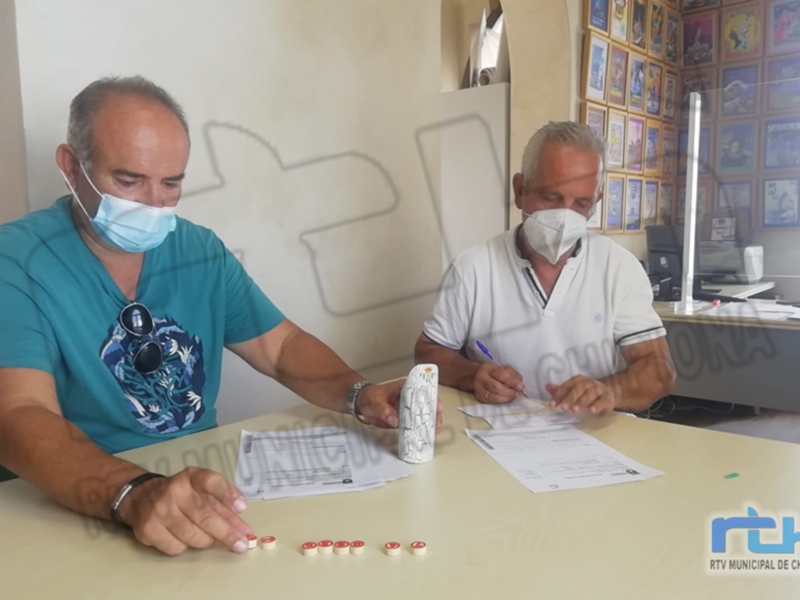 Dos hombres con mascarillas se sientan frente a una mesa, uno de ellos firma un documento mientras el otro observa. La imagen está firmada por RTV Municipal de Chipiona.