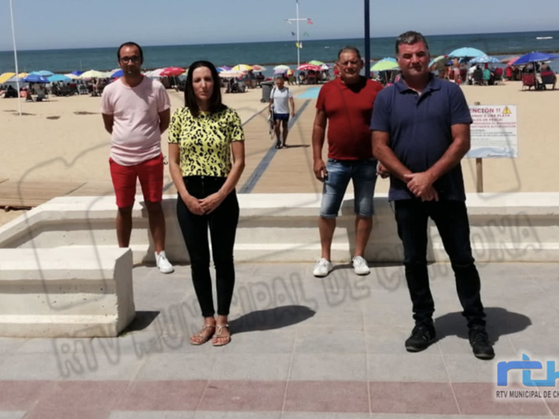 Cuatro personas posan juntas en un muelle frente a una playa con sombrillas y el mar al fondo.