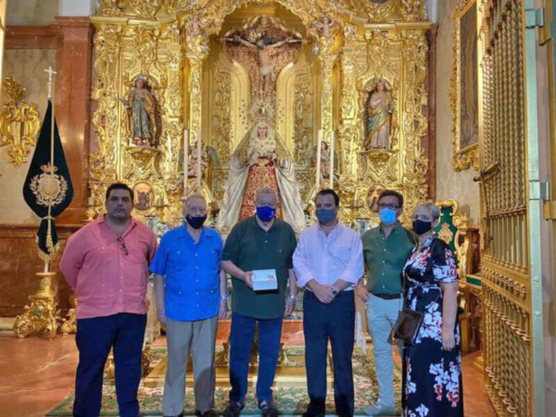 Grupo de personas en una iglesia, posando frente a una imagen religiosa dorada. Todos llevan mascarillas y están en una posición formal, sosteniendo un objeto en la mano central. La imagen sugiere una ceremonia o evento religioso en curso.