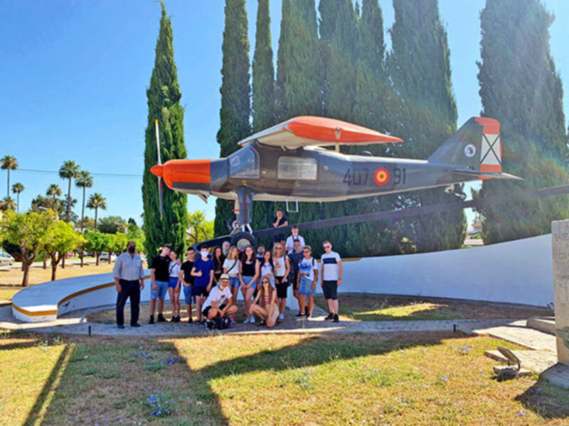 Un grupo de personas posa junto a un avión de combate expuesto al aire libre, rodeado de árboles y bajo un cielo azul claro.