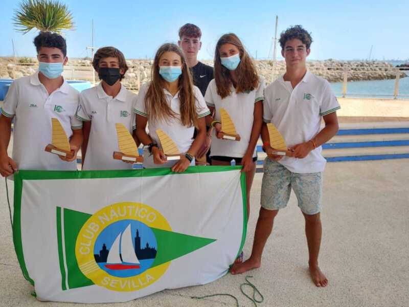 Quatro jovens segurando prêmios e bandeira do Clube Náutico de Sevilha, todos usando máscaras protetoras.