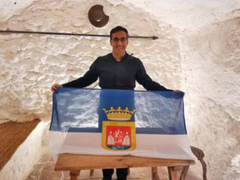 Un hombre sostiene una bandera con un escudo de armas en un interior con paredes de ladrillo y un suelo de baldosas.