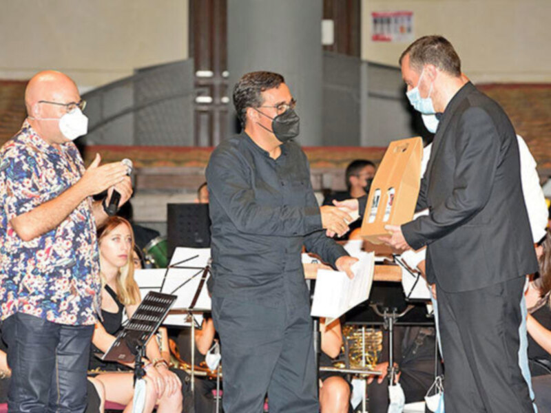 Un hombre en un traje negro entrega un certificado a otro hombre que lleva una camisa con estampado floral. Ambos están en una sala con una orquesta en el fondo.
