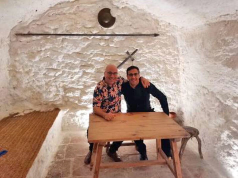 Dos personas sentadas en una mesa de madera en un entorno rural con paredes de arcilla y herramientas de labranza en el fondo.