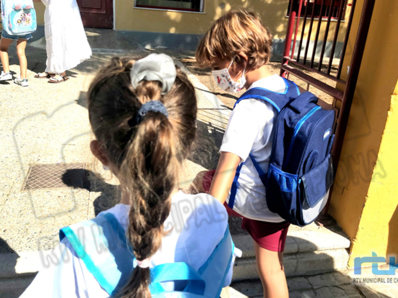 Niños con mochilas en el exterior de una escuela, con una niña con una mochila azul y una niña con una mochila azul y blanca.