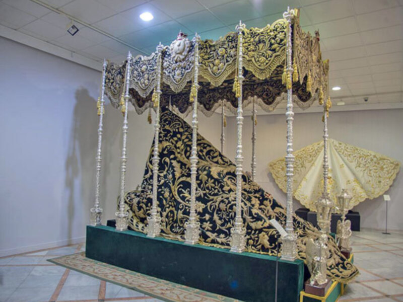 Un hermoso trono de estilo oriental con detalles dorados y negro, rodeado por una alfombra con patrones geométricos, en un ambiente que parece ser una sala de exposiciones o museo.
