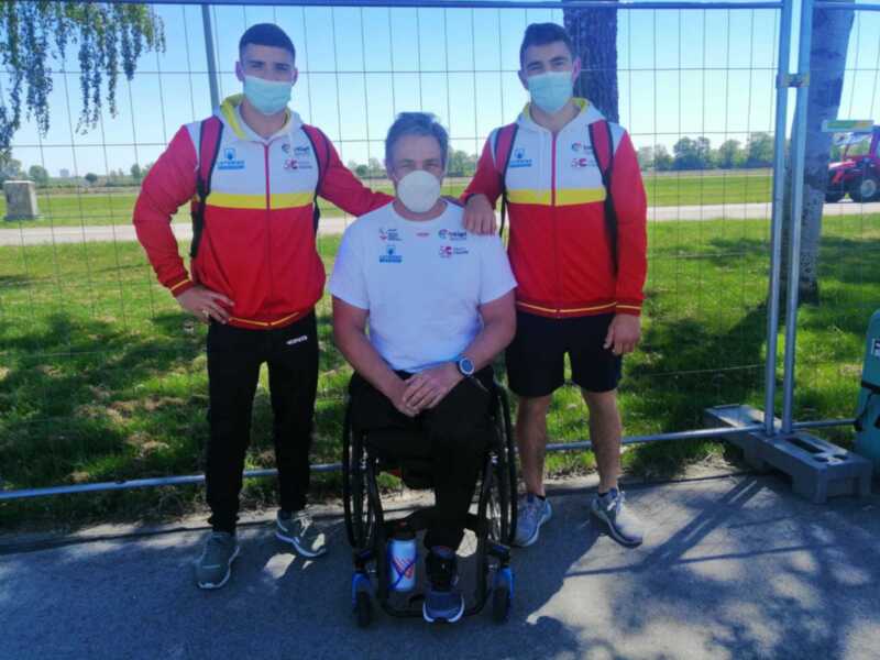 Tres atletas con mascarillas y chaquetas de deporte posan juntos al aire libre, uno en silla de ruedas.