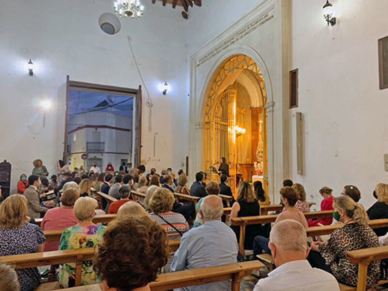 Una multitud de personas asiste a una conferencia o evento en un salón con paredes blancas y una gran ventana. La gente está sentada en bancos de madera, mirando hacia adelante. En la pared hay un cartel con letras en griego y una imagen de una iglesia o templo. La iluminación es natural, proveniente de la ventana y las luces del techo.