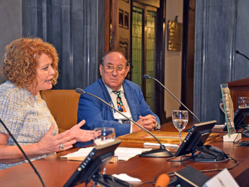 Conferencia de prensa en un entorno formal con dos participantes sentados en una mesa con micrófonos y documentos.
