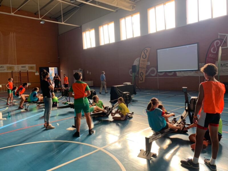 Niños participando en un entrenamiento de remo en un gimnasio con un panel de entrenamiento en el fondo.