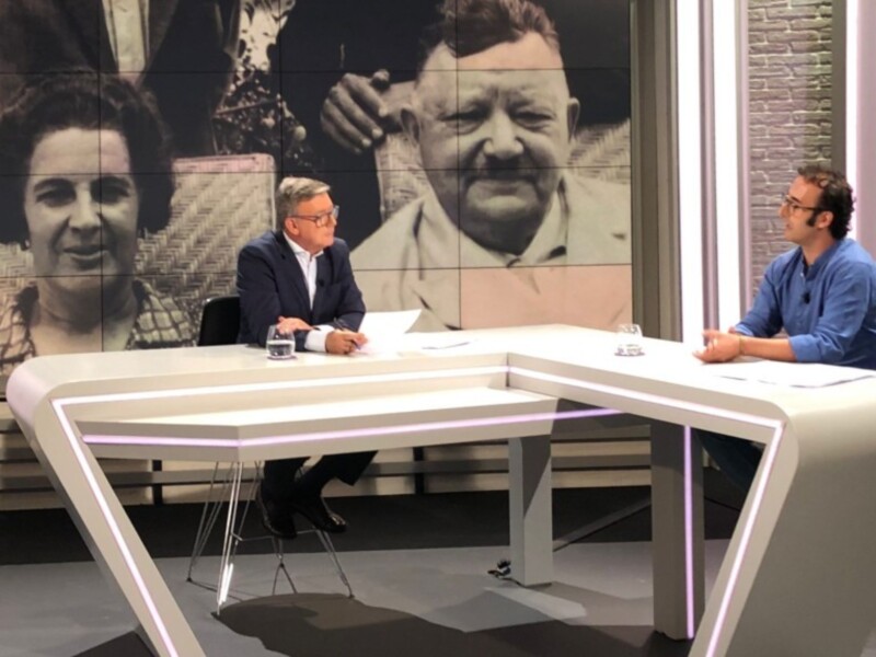 Entrevista en un estudio de televisión con dos personas sentadas en una mesa curva, fondo con retratos de personas históricas.
