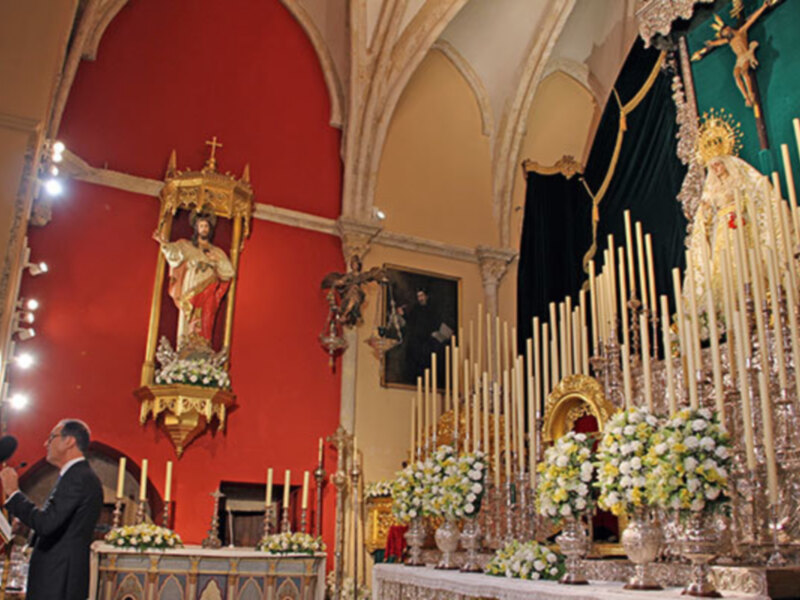Interior de una iglesia con un altar adornado con flores y velas, y una imagen de una figura religiosa en el fondo.