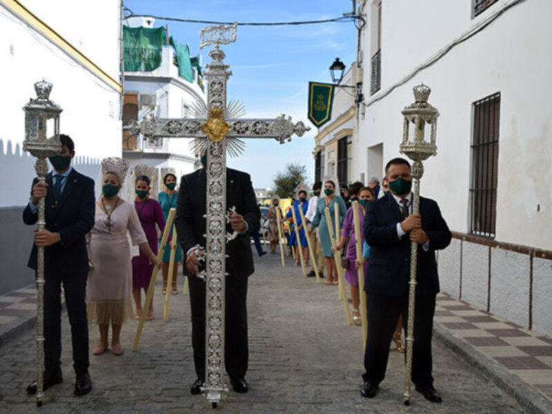 Procesión con portadores de cruces en una calle adoquinada, con edificios blancos y techos verdes en el fondo.
