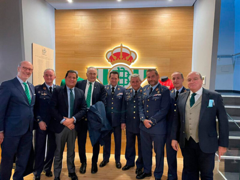 Grupo de personas en uniforme y trajes formales posando frente a un logo de Real Betis Balompié.