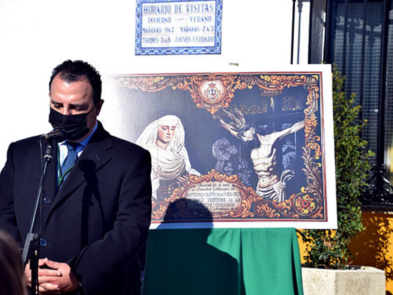 Un hombre en traje negro y máscara negra se encuentra en un escenario, frente a una pancarta con una imagen religiosa y un horario de visitas. El contexto sugiere una ceremonia o evento religioso.
