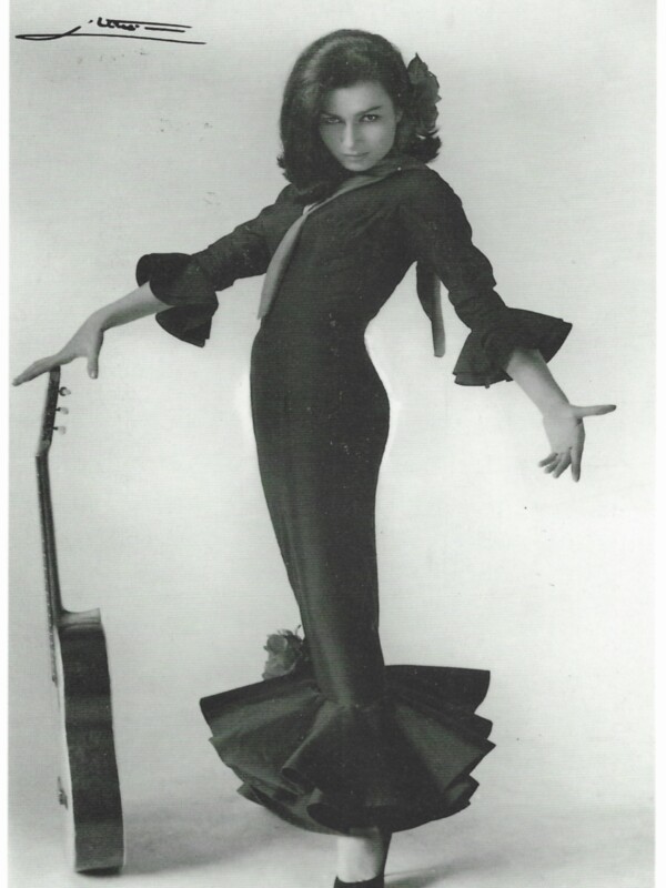 Vintage black and white photo of a woman in a long, elegant dress with a bow in her hair, posing gracefully with one hand on a railing.