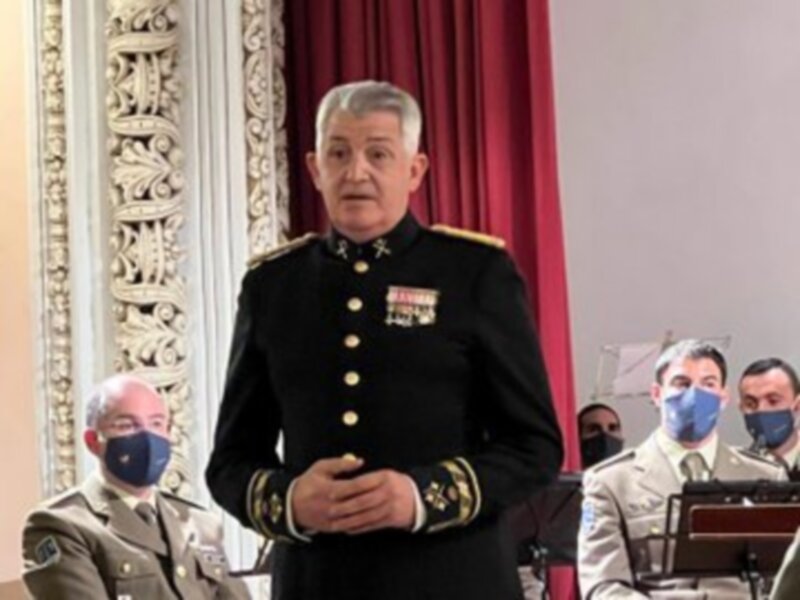 Oficial en uniforme militar hablando en un evento formal, con otros uniformados en el fondo.