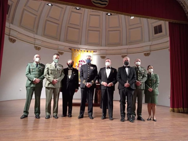 Oficiales militares y civiles en una ceremonia formal en un salón con decoración clásica y máscaras faciales.