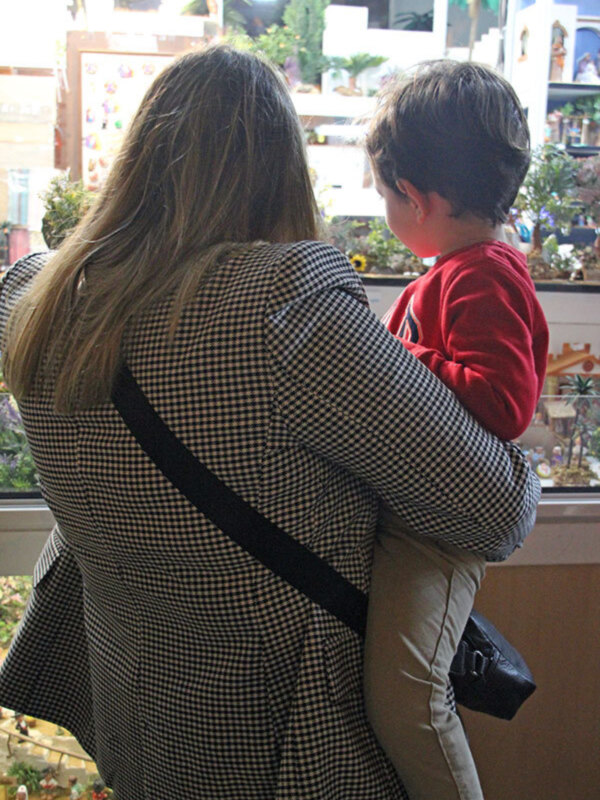 Una madre y su hijo pequeño, ambos de espaldas, frente a una ventana con plantas y decoraciones navideñas. La madre lleva un abrigo a cuadros y el niño, un suéter rojo. La escena transmite una sensación de calidez y familiaridad.