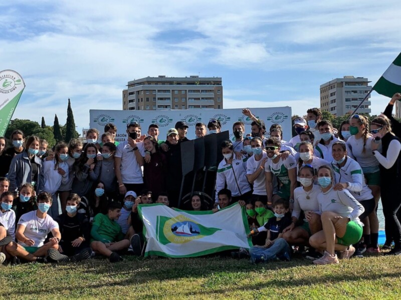 Equipo de remo celebrando junto a su bandera y banner de la Federación Andaluza de Remo en una jornada al aire libre.