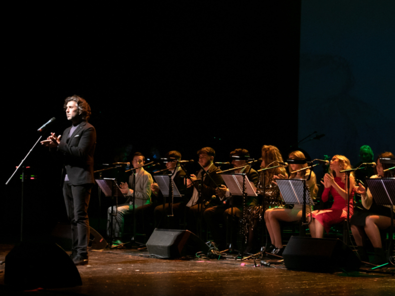 Un intérprete en escena con un coro y orquesta en segundo plano, todos con instrumentos y micrófonos en una presentación musical.