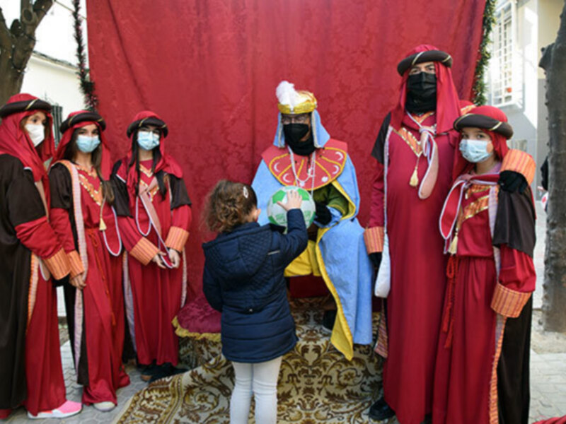 Personas vestidas con trajes tradicionales posan junto a una figura representando a los Reyes Magos durante una celebración en una calle decorada.