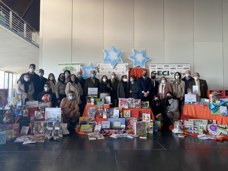 Una representación de personas posando junto a una mesa llena de juguetes y libros, con estrellas azules en el fondo. La escena parece ser una donación o una presentación de regalos.