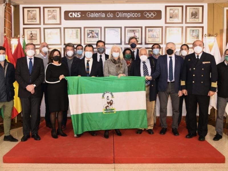 Grupo de personas en una reunión formal con máscaras, sosteniendo una bandera de Andalucía y fondo con carteles de olímpicos.