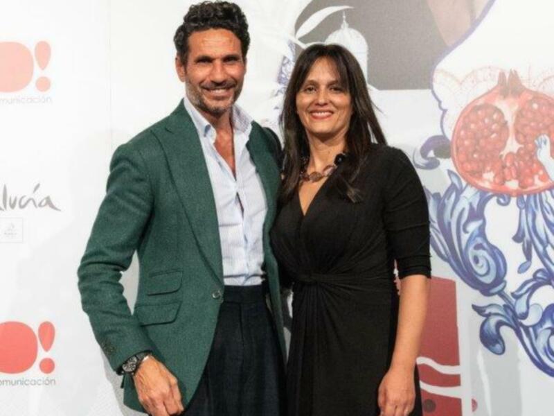 Un hombre y una mujer posan frente a un fondo con un diseño floral y una fruta roja, posiblemente en una celebración o evento.