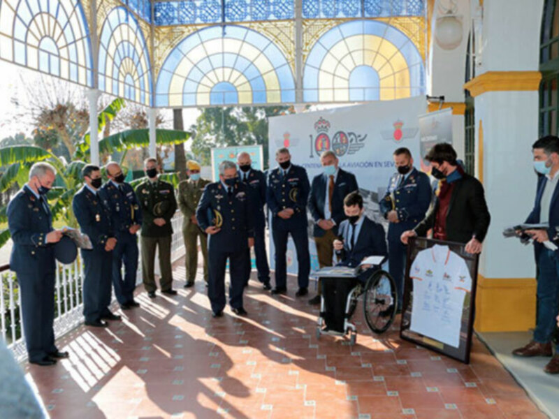 Personalidades militares y civiles en una ceremonia formal, con un hombre en silla de ruedas en el centro, en un entorno con ventanas arqueadas y decoración floral. El evento parece ser un acto de reconocimiento o premio.